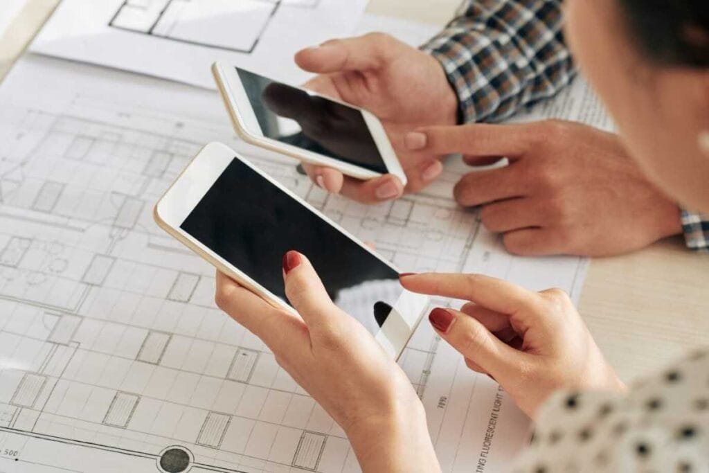 Two people using white smartphones while discussing plans on a blueprint.